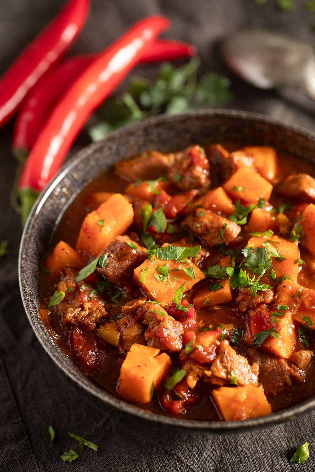 bowl of stew with pork neck pieces and sweet potatoes, a few long red chilies and a spoon near the bowl.