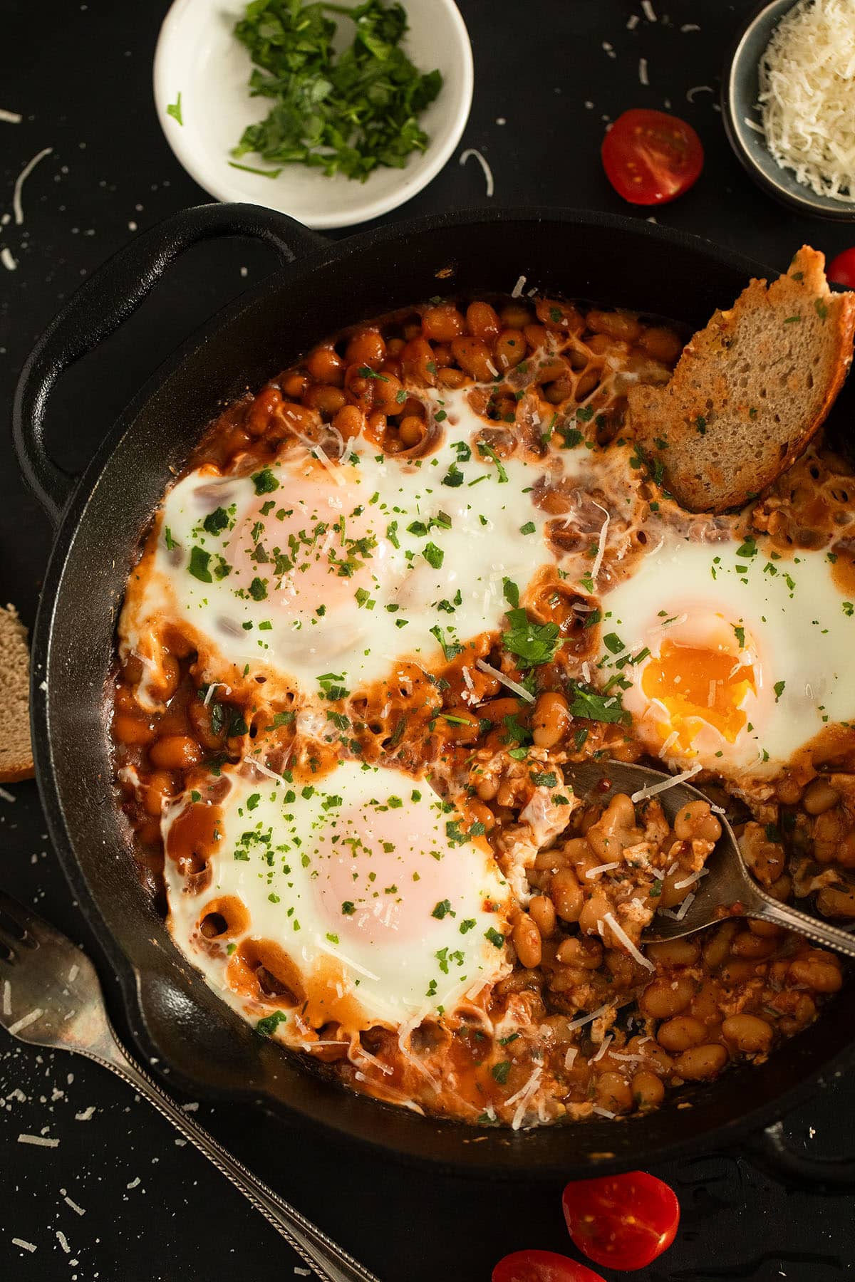 a skillet with baked beans and eggs, a slice of bread dipped into it.