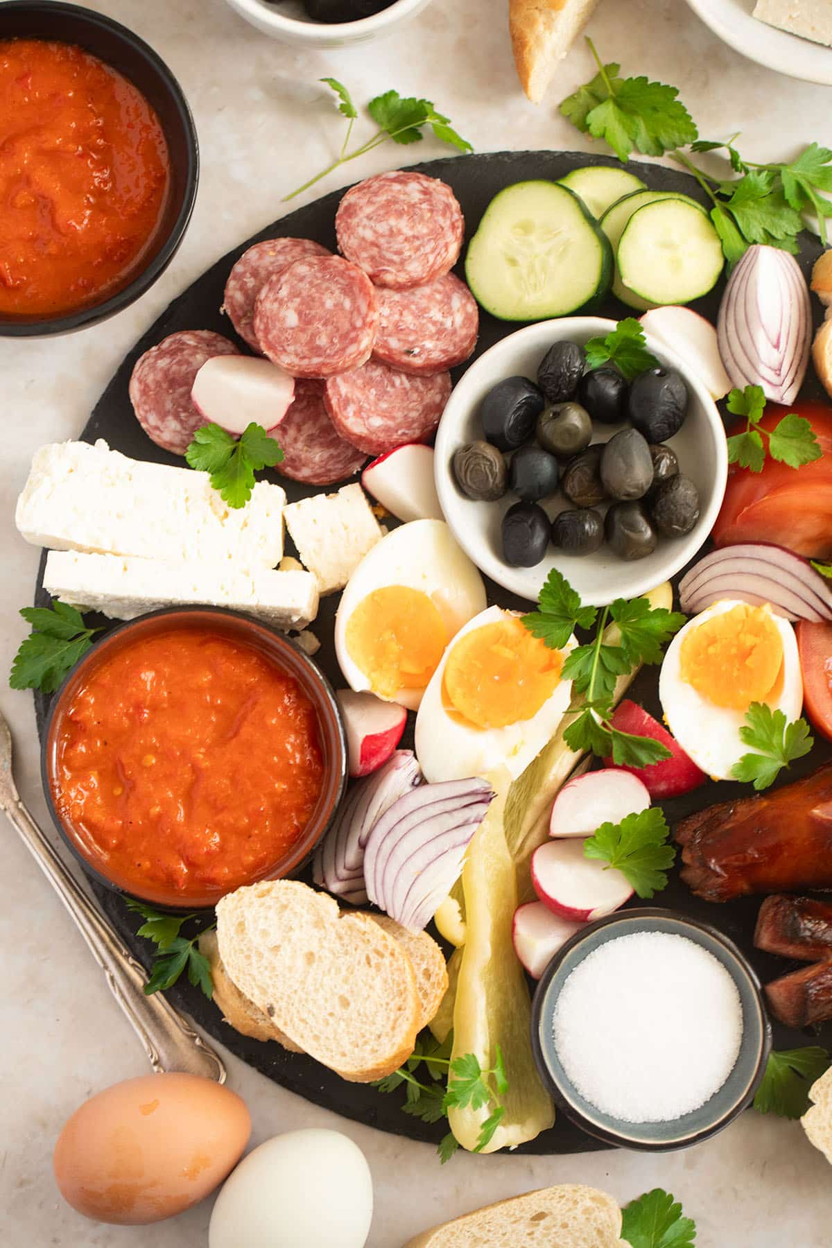 a large platter with balkan breakfast with bread, eggs, feta, sausage, vegetables, and olives.