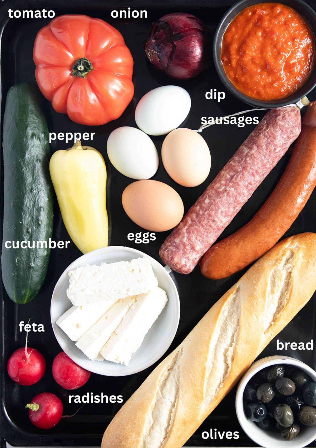 labeled ingredients for making balkan breakfast: bread, tomato, dip, onion, eggs, pepper, cucumber, feta, radishes, olives, sausages.