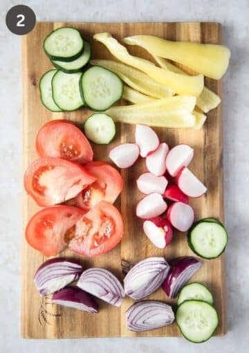 sliced cucumbers, peppers, tomatoes, radishes, and onions on a wooden board.