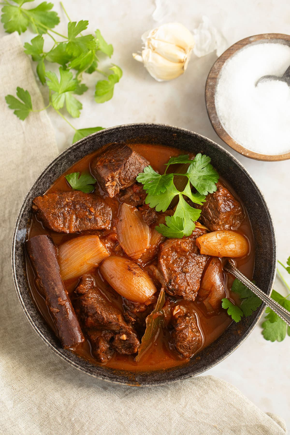bowl of lamb stifado garnished with parsley leaves, more parsley, salt and garlic beside it.