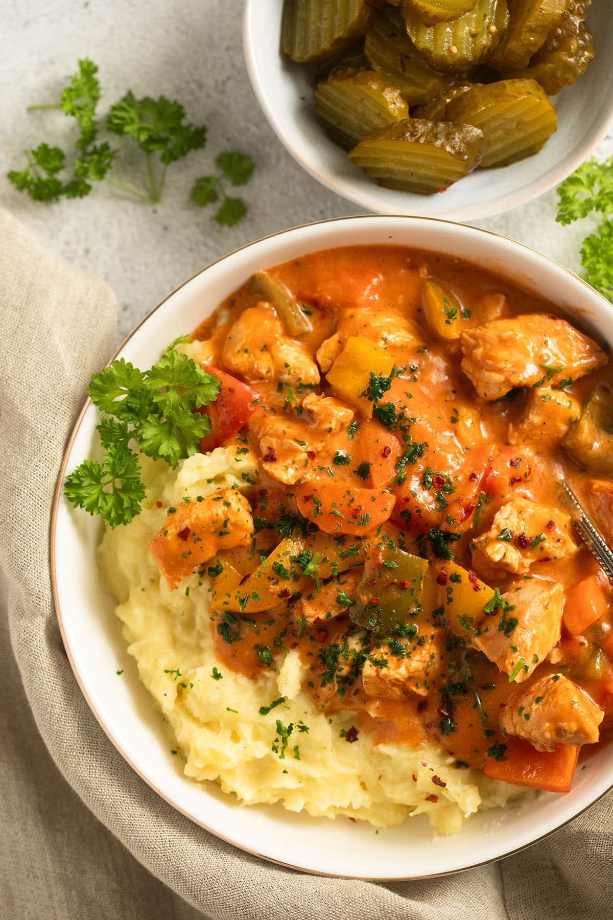 a bowl with goulash made with chicken, peppers and lots of sauce and a bowl of pickles.