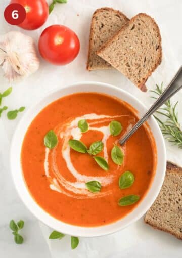 bowl of roasted tomato and garlic soup, slices of bread, two tomatoes, basil, and a bulb of garlic.