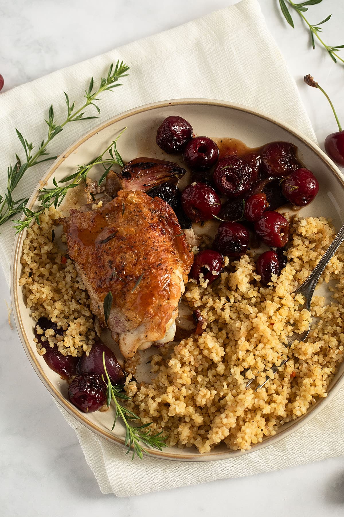 a plate with a crispy chicken thighs, cherry sauce, cooked bulgur, and thyme sprigs.