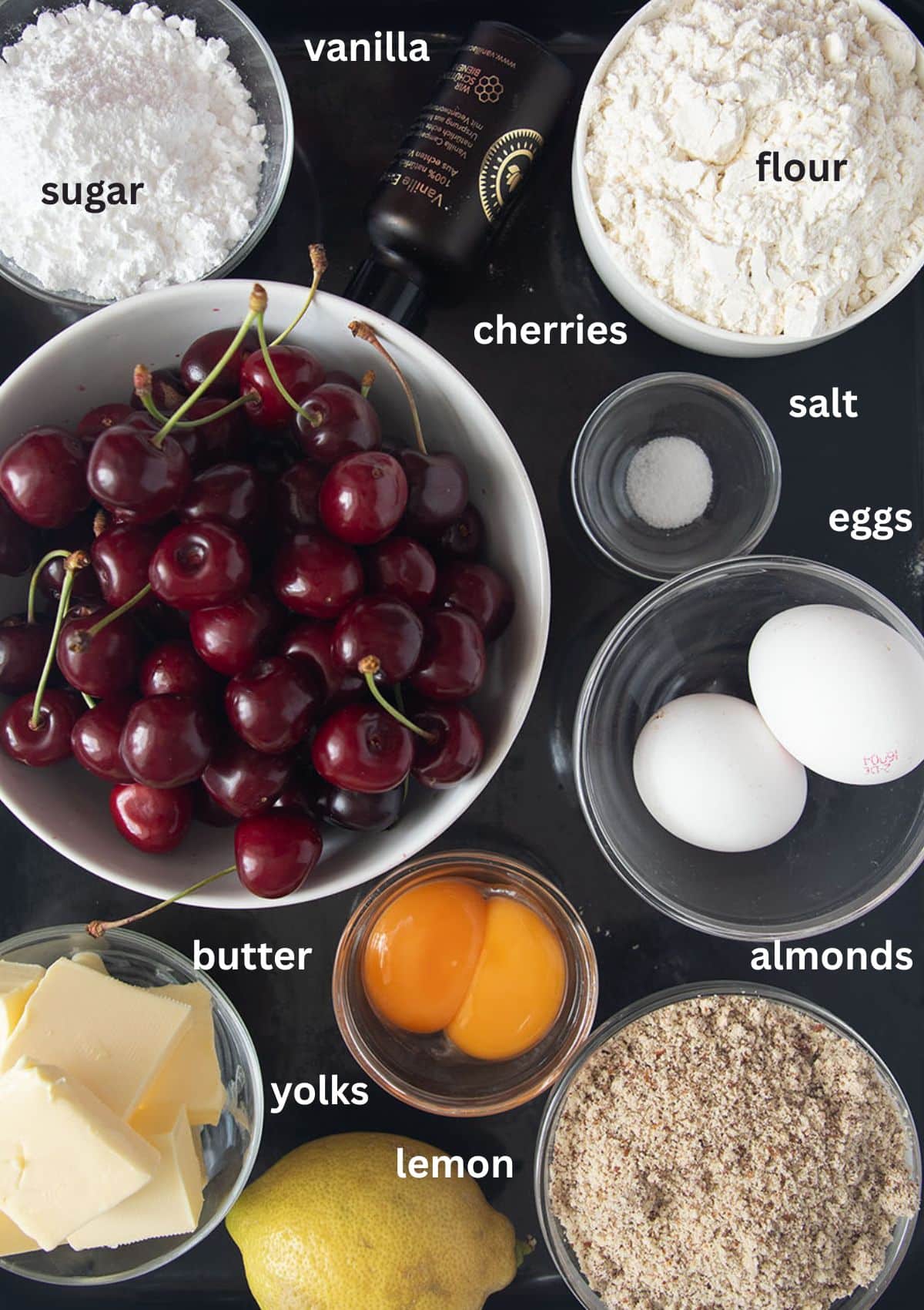 labeled ingredients fo rmaking cherry tart with fresh cherries, almonds, lemon, sugar, eggs, and flour.