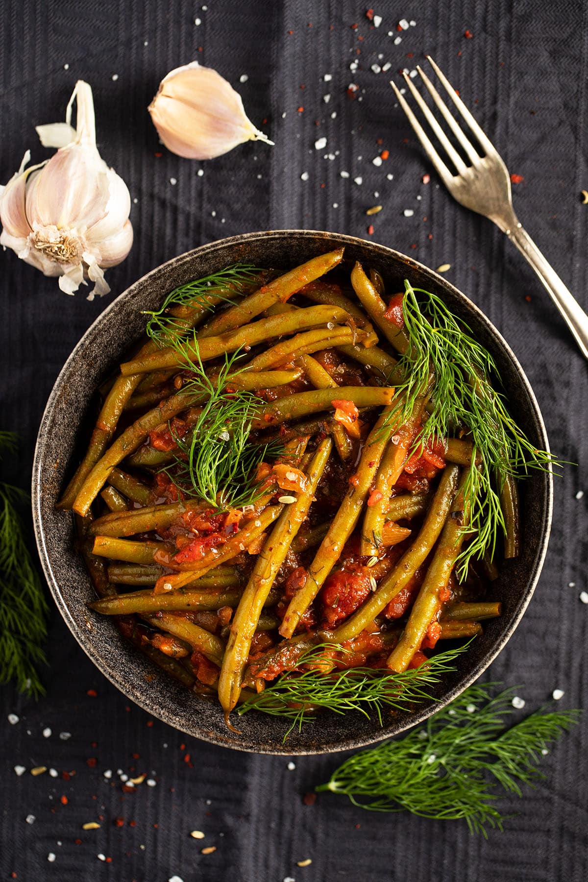 one plate with green bean stew, garlic bulbs and a fork on a table.
