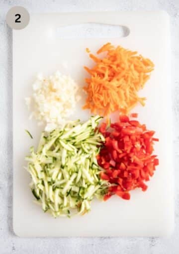 grated carrots and zucchini, finely chopped onions and peppers on a cutting board.
