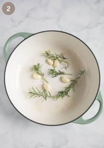 large garlic cloves and rosemary sprigs in a dutch oven ready for cooking duck legs.
