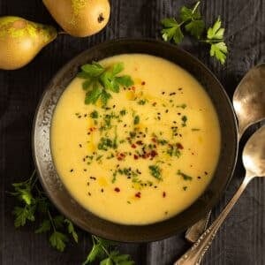 parsnip and potato soup in a bowl, two spoons, fresh parsley, and two pears around it.