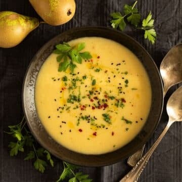 parsnip and potato soup in a bowl, two spoons, fresh parsley, and two pears around it.