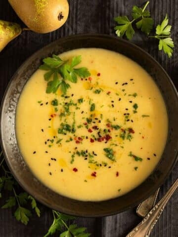 parsnip and potato soup in a bowl, two spoons, fresh parsley, and two pears around it.