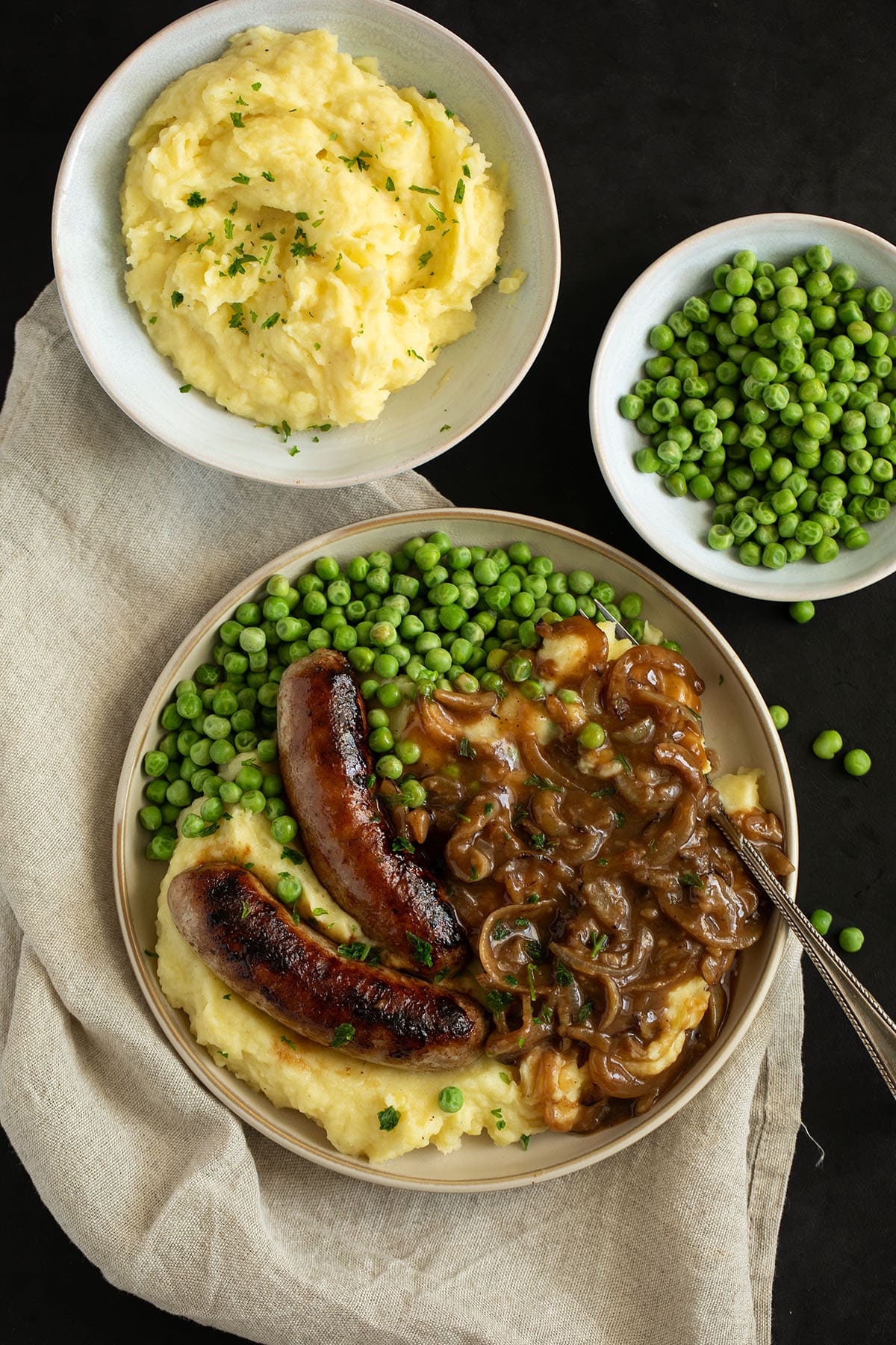 one plate with bangers, mash, and peas and two small bowls with mashed potatoes and cooked green peas.