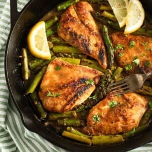 four pieces of golden chicken breasts with asparagus and lemon slices in a skillet.