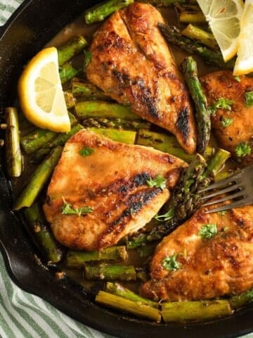 four pieces of golden chicken breasts with asparagus and lemon slices in a skillet.