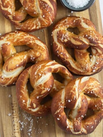 many german pretzels sprinkled with coarse salt on a wooden cutting board.