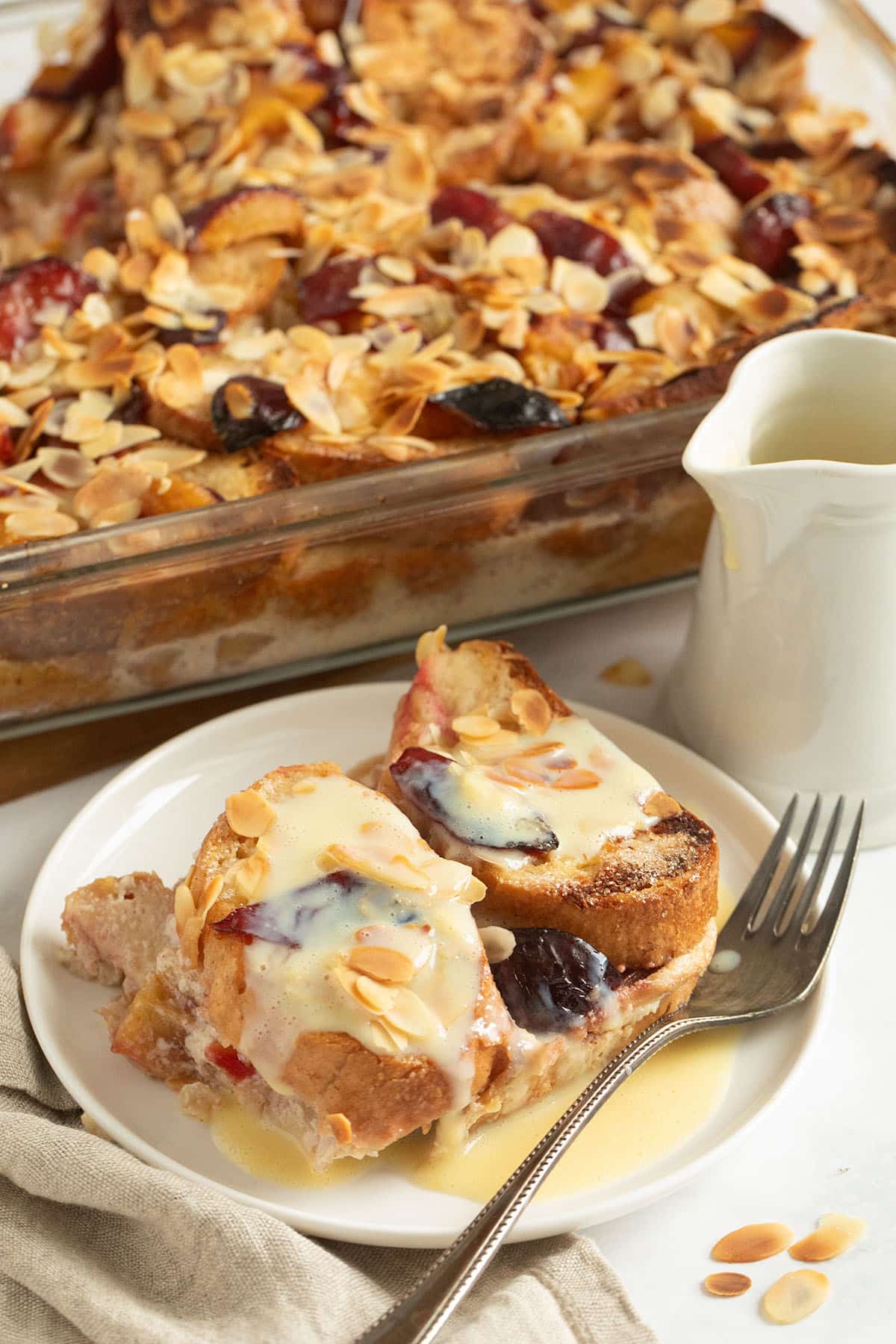 plum bread pudding covered with homemade vanilla sauce on a plate, the rest of the pudding behind it.